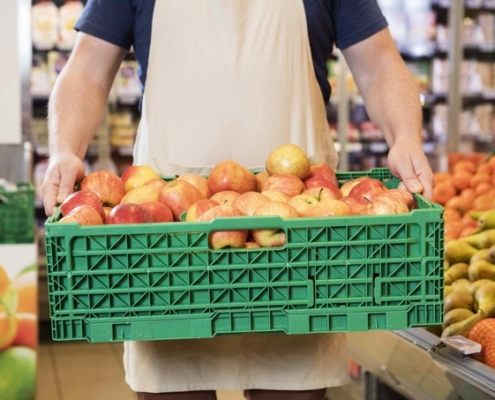 A grocery retail employee carries a crate of apples to the produce department
