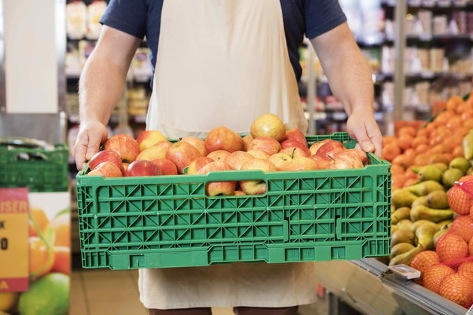 A grocery retail employee carries a crate of apples to the produce department